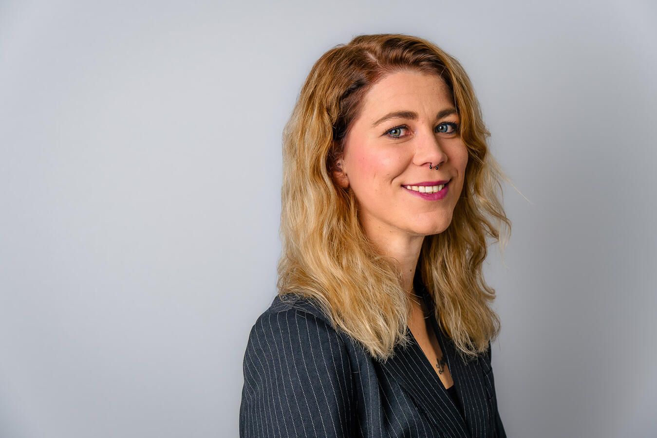 Ein natürliches Portrait einer jungen Frau mit lockigem blondem Haar vor neutralem Hintergrund. Das Bild zeigt einen freundlichen, authentischen Ausdruck und wurde mit weichem, professionellem Licht aufgenommen – ideal für Business- oder Lifestyle-Darstell
